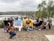 A group of smiling individuals poses together near a serene lake, holding banners with 'Coastal Cleanup Day' and 'World Cleanup Day' written on them. The participants are dressed casually and some hold cleaning tools and buckets, indicating their involvement in an environmental cleanup activity. The background features a calm body of water surrounded by trees and hills under an overcast sky.