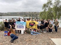 A group of smiling individuals poses together near a serene lake, holding banners with 'Coastal Cleanup Day' and 'World Cleanup Day' written on them. The participants are dressed casually and some hold cleaning tools and buckets, indicating their involvement in an environmental cleanup activity. The background features a calm body of water surrounded by trees and hills under an overcast sky.