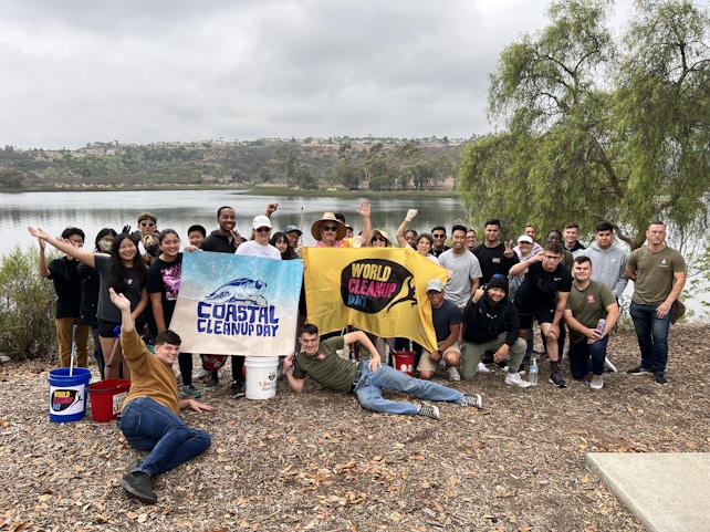 A group of smiling individuals poses together near a serene lake, holding banners with 'Coastal Cleanup Day' and 'World Cleanup Day' written on them. The participants are dressed casually and some hold cleaning tools and buckets, indicating their involvement in an environmental cleanup activity. The background features a calm body of water surrounded by trees and hills under an overcast sky.