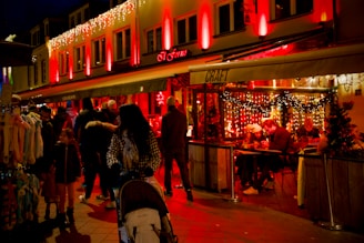 A lively street scene featuring a warmly lit restaurant adorned with festive lights. Several people are gathered around tables inside the restaurant, enjoying food and conversation. Outside, a diverse group of people, including a woman pushing a stroller and several shoppers, walk by illuminated shops. The ambiance is cozy and inviting, enhanced by the red and yellow lighting.