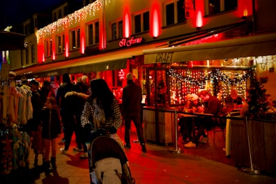 A lively street scene featuring a warmly lit restaurant adorned with festive lights. Several people are gathered around tables inside the restaurant, enjoying food and conversation. Outside, a diverse group of people, including a woman pushing a stroller and several shoppers, walk by illuminated shops. The ambiance is cozy and inviting, enhanced by the red and yellow lighting.