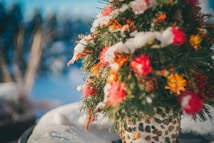 A decorative arrangement featuring colorful flowers interspersed with sprigs of pine, all dusted with snow, set within a textured vase made of stones. The background is a softly blurred winter landscape.