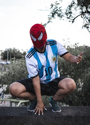 A person wearing a red Spider-Man mask and an Argentina soccer jersey squats on a ledge in an outdoor setting. The jersey features light blue and white stripes with the number 10. Surrounding foliage and a slightly overcast sky are visible in the background.