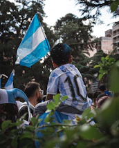 A child in an Argentina soccer jersey sitting on shoulders holding a blue flag