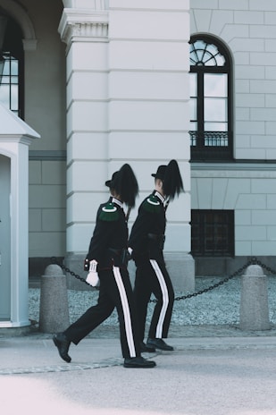 Two uniformed guards in black attire with distinctive white stripes and feathered hats are marching in synchronization along a building with arched windows and classical architecture.
