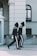 Two uniformed guards in black attire with distinctive white stripes and feathered hats are marching in synchronization along a building with arched windows and classical architecture.