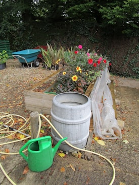 A garden scene with a wooden planter box filled with various colorful flowers, including pink and yellow blooms. A gray barrel and a green watering can are on the ground beside a coiled yellow hose. A wheelbarrow is positioned in the background next to several potted plants. Leaves are scattered on the ground, giving a sense of an outdoor space surrounded by foliage.