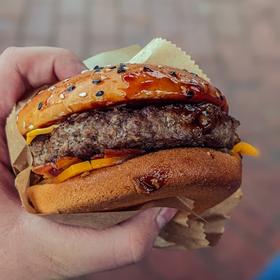 A close-up of a freshly prepared burger held in someone's hand, featuring a sesame seed bun, a juicy beef patty, melted cheese, and a sauce glaze.