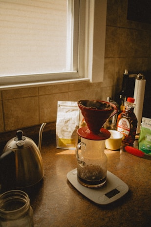 A cozy kitchen scene with a window providing soft natural light. A red ceramic coffee dripper sits atop a glass carafe on a digital scale. A gooseneck kettle is nearby, with various coffee products such as beans and syrup visible in the background.