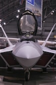A sleek, modern fighter jet with a sharp, aerodynamic design is positioned inside a spacious hangar. The cockpit canopy is open, revealing part of the interior. Overhead lighting casts a soft glow on the aircraft's smooth surface, highlighting its stealthy angles and engineering.
