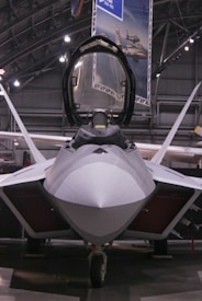 A sleek, modern fighter jet with a sharp, aerodynamic design is positioned inside a spacious hangar. The cockpit canopy is open, revealing part of the interior. Overhead lighting casts a soft glow on the aircraft's smooth surface, highlighting its stealthy angles and engineering.