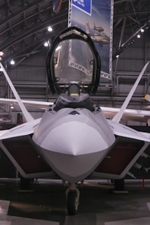 A sleek, modern fighter jet with a sharp, aerodynamic design is positioned inside a spacious hangar. The cockpit canopy is open, revealing part of the interior. Overhead lighting casts a soft glow on the aircraft's smooth surface, highlighting its stealthy angles and engineering.