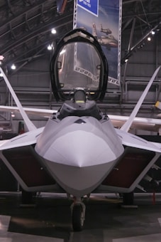A sleek, modern fighter jet with a sharp, aerodynamic design is positioned inside a spacious hangar. The cockpit canopy is open, revealing part of the interior. Overhead lighting casts a soft glow on the aircraft's smooth surface, highlighting its stealthy angles and engineering.
