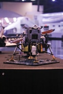 A detailed Lego model of a lunar module with an astronaut figurine standing on a base that resembles the moon's surface. The model features an American flag and is built with intricate parts to mimic a real spacecraft. It's displayed indoors with what seems to be a museum or exhibition setting in the background, featuring blurred lights and other structures.