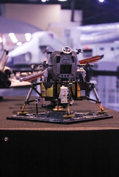 A detailed Lego model of a lunar module with an astronaut figurine standing on a base that resembles the moon's surface. The model features an American flag and is built with intricate parts to mimic a real spacecraft. It's displayed indoors with what seems to be a museum or exhibition setting in the background, featuring blurred lights and other structures.