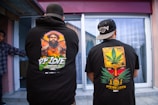 Two individuals are standing with their backs facing the camera, wearing black hoodies with bold, colorful graphics on the back. One hoodie features a portrait of a man surrounded by vibrant colors and the words 'PF Love'. The other hoodie displays a graphic of a face partially covered by a large cannabis leaf and the text 'Steven Loco'. Both individuals wear black caps turned backward.