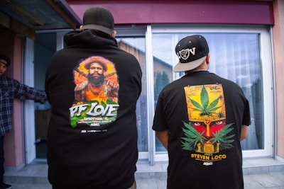 Two individuals are standing with their backs facing the camera, wearing black hoodies with bold, colorful graphics on the back. One hoodie features a portrait of a man surrounded by vibrant colors and the words 'PF Love'. The other hoodie displays a graphic of a face partially covered by a large cannabis leaf and the text 'Steven Loco'. Both individuals wear black caps turned backward.