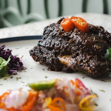 A richly spiced, grilled piece of meat topped with sliced red chili peppers. The dish is paired with vibrant garnishes including purple cabbage and shredded vegetables, providing a colorful contrast on a white plate.