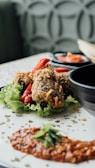 Crispy fried chicken pieces garnished with red chili slices rest on a bed of fresh green lettuce. A dollop of red sauce with a garnish of herbs is plated nearby, and a black bowl is partially visible in the background. The table surface has a light color with a subtle pattern.