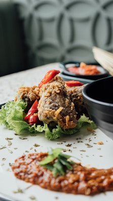 Crispy fried chicken pieces garnished with red chili slices rest on a bed of fresh green lettuce. A dollop of red sauce with a garnish of herbs is plated nearby, and a black bowl is partially visible in the background. The table surface has a light color with a subtle pattern.