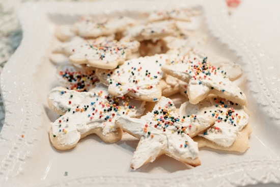 A collection of star-shaped cookies is arranged on a decorative white plate. The cookies are topped with white frosting and sprinkled with small, colorful candy sprinkles. The plate is intricately designed with embossed patterns around the edge.