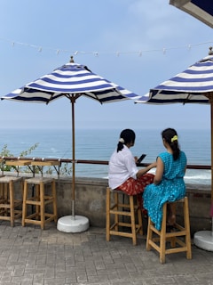 Two people are sitting on wooden stools under striped umbrellas at an outdoor cafe overlooking the ocean. They appear to be engaged in conversation, with one holding a phone. The ocean is calm and the sky is clear with a string of lights above them.