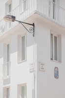 A corner of a white building features several windows with minimalist railings. Mounted on the wall is a street lamp attached to an ornate metal bracket, and there are plaques with street names 'Travessa da Correios da Ribeira' and 'Travessa do Rosário'. A decorative ceramic tile is embedded in the wall.