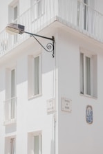 A corner of a white building features several windows with minimalist railings. Mounted on the wall is a street lamp attached to an ornate metal bracket, and there are plaques with street names 'Travessa da Correios da Ribeira' and 'Travessa do Rosário'. A decorative ceramic tile is embedded in the wall.