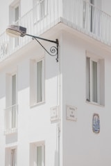 A corner of a white building features several windows with minimalist railings. Mounted on the wall is a street lamp attached to an ornate metal bracket, and there are plaques with street names 'Travessa da Correios da Ribeira' and 'Travessa do Rosário'. A decorative ceramic tile is embedded in the wall.