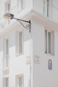 A corner of a white building features several windows with minimalist railings. Mounted on the wall is a street lamp attached to an ornate metal bracket, and there are plaques with street names 'Travessa da Correios da Ribeira' and 'Travessa do Rosário'. A decorative ceramic tile is embedded in the wall.