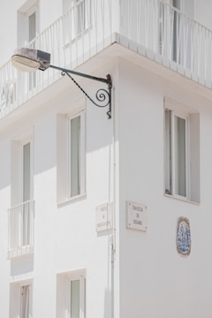 A corner of a white building features several windows with minimalist railings. Mounted on the wall is a street lamp attached to an ornate metal bracket, and there are plaques with street names 'Travessa da Correios da Ribeira' and 'Travessa do Rosário'. A decorative ceramic tile is embedded in the wall.