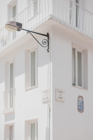 A corner of a white building features several windows with minimalist railings. Mounted on the wall is a street lamp attached to an ornate metal bracket, and there are plaques with street names 'Travessa da Correios da Ribeira' and 'Travessa do Rosário'. A decorative ceramic tile is embedded in the wall.