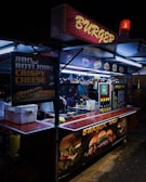 A street food stall selling burgers, brightly lit with neon signs. A vendor is preparing food inside the stall, with various posters showing burger options and condiments on display.