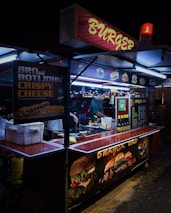 A street food stall selling burgers, brightly lit with neon signs. A vendor is preparing food inside the stall, with various posters showing burger options and condiments on display.