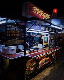 A street food stall selling burgers, brightly lit with neon signs. A vendor is preparing food inside the stall, with various posters showing burger options and condiments on display.