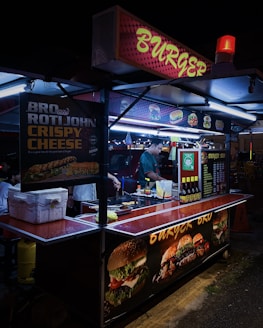 A street food stall selling burgers, brightly lit with neon signs. A vendor is preparing food inside the stall, with various posters showing burger options and condiments on display.