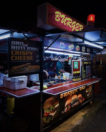 A street food stall selling burgers, brightly lit with neon signs. A vendor is preparing food inside the stall, with various posters showing burger options and condiments on display.
