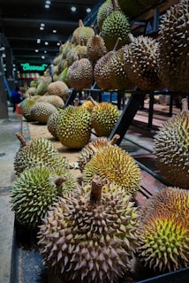 An assortment of durians with spiky shells arranged on the shelves of a market or store, with several rows extending into the background. The lighting highlights the texture and varied coloration of the fruit skins.