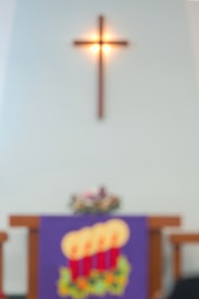 blurred image of a church interior with a wooden crucifix mounted on a wall, above an altar adorned with a purple cloth featuring a design of four glowing candles and some floral decoration.