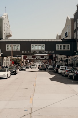 A busy street scene with multiple cars parked alongside the road. An overhead pedestrian bridge connects two buildings with signs reading 'The Clement Monterey' and 'Monterey Canning Co.'. The buildings are multi-story, with various architectural features.