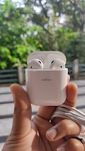 A hand is holding a white Joyroom wireless earphone case with two earphones inside. In the background, there are blurred green trees and a fence, indicating an outdoor setting.