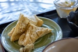 Crispy, golden-brown deep-fried tofu triangles are arranged on a green plate. In the background, there are a few other dishes and cups, suggesting a dining table setup.