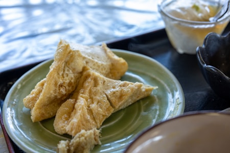 Crispy, golden-brown deep-fried tofu triangles are arranged on a green plate. In the background, there are a few other dishes and cups, suggesting a dining table setup.