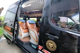 A luxury van with its side door open, revealing a well-appointed interior. The seating is covered with a combination of brown and beige leather, featuring a sophisticated design. Ambient lighting is visible on the ceiling, and multiple large windows allow for natural light.