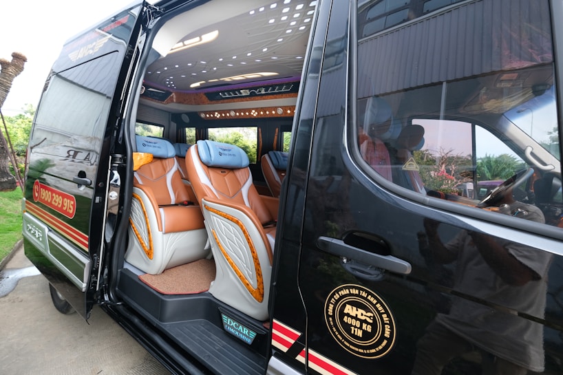 A luxury van with its side door open, revealing a well-appointed interior. The seating is covered with a combination of brown and beige leather, featuring a sophisticated design. Ambient lighting is visible on the ceiling, and multiple large windows allow for natural light.