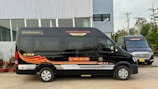 Two black vans are parked in front of a modern industrial building. The vans have red and gold designs on their sides, along with text and a phone number. The ground is paved, and there are some potted plants lined up near the building. Power lines and utility poles are visible in the background.