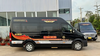 Two black vans are parked in front of a modern industrial building. The vans have red and gold designs on their sides, along with text and a phone number. The ground is paved, and there are some potted plants lined up near the building. Power lines and utility poles are visible in the background.