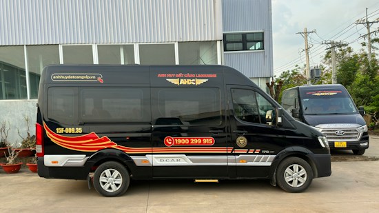 Two black vans are parked in front of a modern industrial building. The vans have red and gold designs on their sides, along with text and a phone number. The ground is paved, and there are some potted plants lined up near the building. Power lines and utility poles are visible in the background.