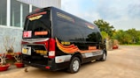 A black minibus with red and yellow graphics is parked outside a building with large windows. The vehicle has contact information and logos, and there are decorative plants in red pots nearby. In the background, there are trees and a clear sky.
