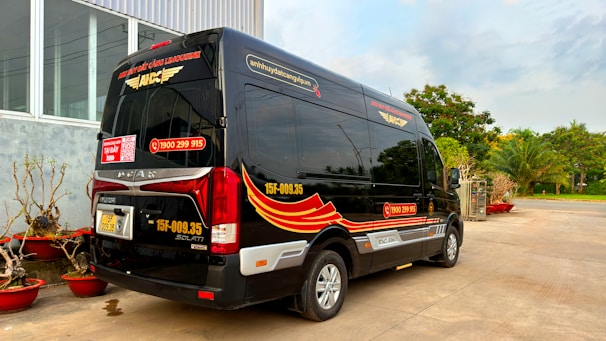 A black minibus with red and yellow graphics is parked outside a building with large windows. The vehicle has contact information and logos, and there are decorative plants in red pots nearby. In the background, there are trees and a clear sky.